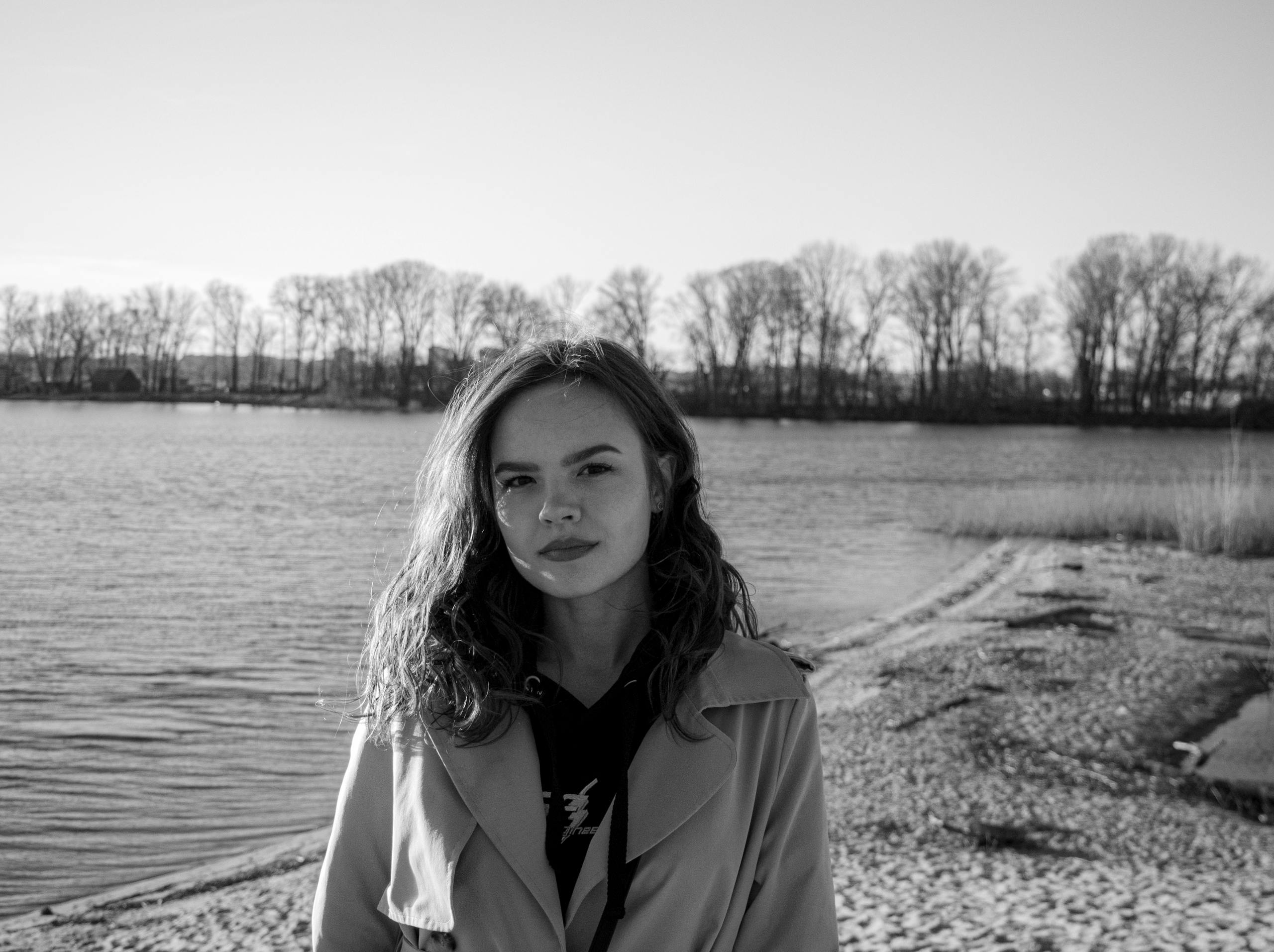 Monochrome portrait of a woman posing near a serene lakeside with trees in the background.